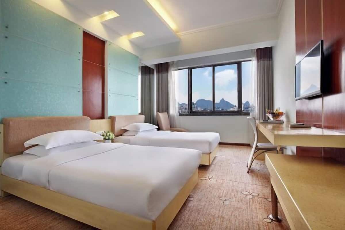 A hotel room with two beds, a desk, a TV, and a large window with a city view.