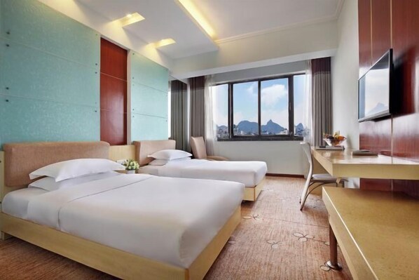 Quarto de hotel com duas camas, uma escrivaninha, televisão e uma janela grande com vista para a cidade.
