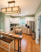 Una cocina con armarios blancos, un refrigerador de acero inoxidable, una mesa de comedor de madera y una lámpara colgante.