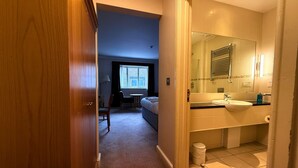 A hotel room with a bed, a desk, a chair, a window with curtains, a bathroom with a sink and mirror, and a trash can. - Relaxing Executive Double Room with Sofa in Blaby (Blaby)