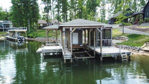 Un muelle de madera con un área de descanso cubierta y un pequeño edificio, rodeado de árboles y un cuerpo de agua.