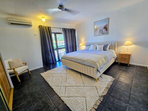 Ein Schlafzimmer mit Bett, Sessel, Nachttisch, Lampe, Klimaanlage, Deckenventilator, einer Tür mit Vorhängen und einem Bild an der Wand.