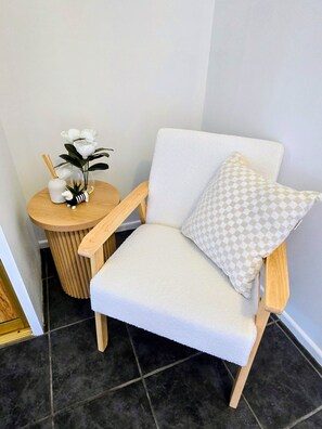 Ein weißer Sessel mit kariertem Kissen, ein kleiner Holztisch mit einer Blumenvase und einem Schilfdiffusor.