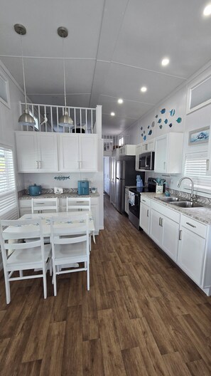 A modern kitchen with white cabinets, a wooden floor, and a dining area with white chairs. - Happier in the keys- waterfront tiny home in Key Largo (Key Largo)