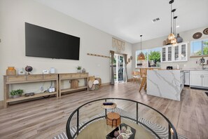 A spacious living area with a large flat-screen TV, wooden shelving, a glass coffee table, and a kitchen with white cabinets and a marble countertop. - Elegant Mediterranean 3BR Haven Citrus Heights (Citrus Heights)