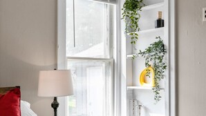 A bedroom with a bed, a nightstand, a lamp, a window with blinds, and a shelf with plants. - Roomy Central Hub • Games, Fire Pit & Downtown Fun (Oshkosh)