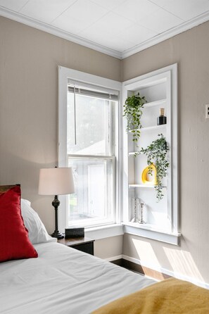 Une chambre à coucher comprenant un lit, une table de chevet, une lampe, une fenêtre avec des stores et une étagère avec des plantes.