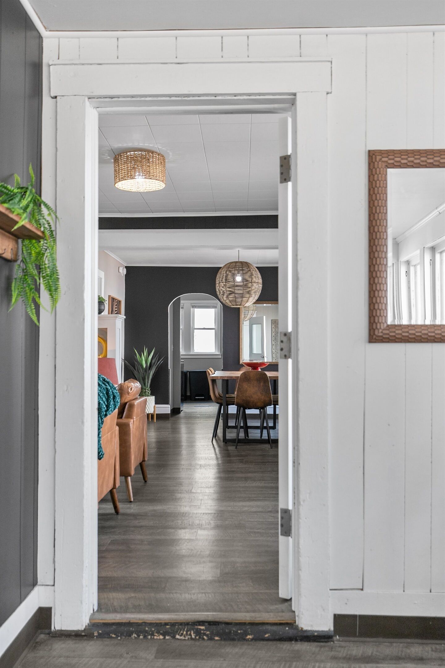 A hallway leading to a dining area with a wooden floor, a hanging light fixture, and a mirror on the wall.