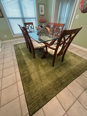 A dining area with a glass-top table surrounded by wooden chairs, a green rug, and a window with blinds. - Sunsets! Beachfront!  Gulf view! 2-bedroom condo in beautiful Perdido Key, FL (Pensacola)