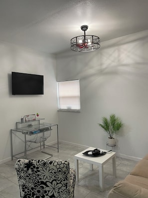 Ein Wohnzimmer mit Flachbildfernseher, Deckenventilator, einem kleinen Tisch mit Pflanze, einem gemusterten Sessel und einem weißen Sofa.