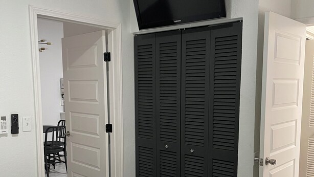 A bedroom with a bed, a wardrobe, a television mounted above the wardrobe, and a door leading to another room.