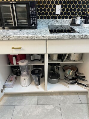 Una cucina con un forno a microonde nero, un frullatore bianco e diverse pentole e padelle sugli scaffali.