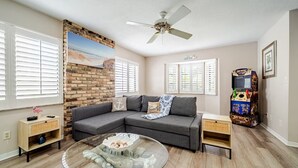A living room with a sectional sofa, a glass coffee table, a brick accent wall, a ceiling fan, and a claw machine.