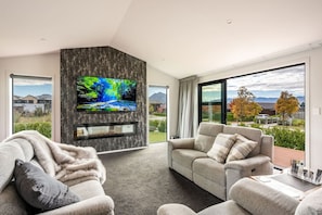 A modern living room with a flat-screen TV mounted on a textured wall, a grey sofa, and a large window offering a view of the outdoors.