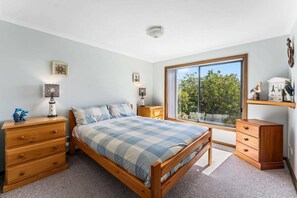 Ein Schlafzimmer mit Bett, Holzkommode, Nachttischen, einem Fenster mit Ausblick und Wanddekoration.