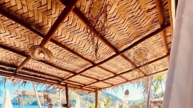 Uma área externa coberta com piso de madeira, móveis de vime e vista para a praia.