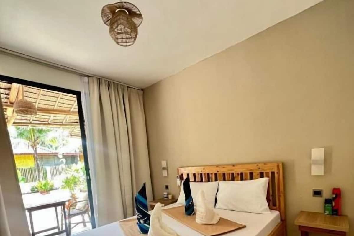 Ein hölzernes Bett mit weißen Leinen, ein kleiner Nachttisch und ein Blick ins Freie durch eine Glastür.