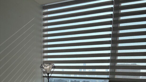 Ein modernes Wohnzimmer mit einer Couch, Sesseln, einem Glastisch und Blick auf eine Stadtlandschaft durch große Fenster mit Jalousien.