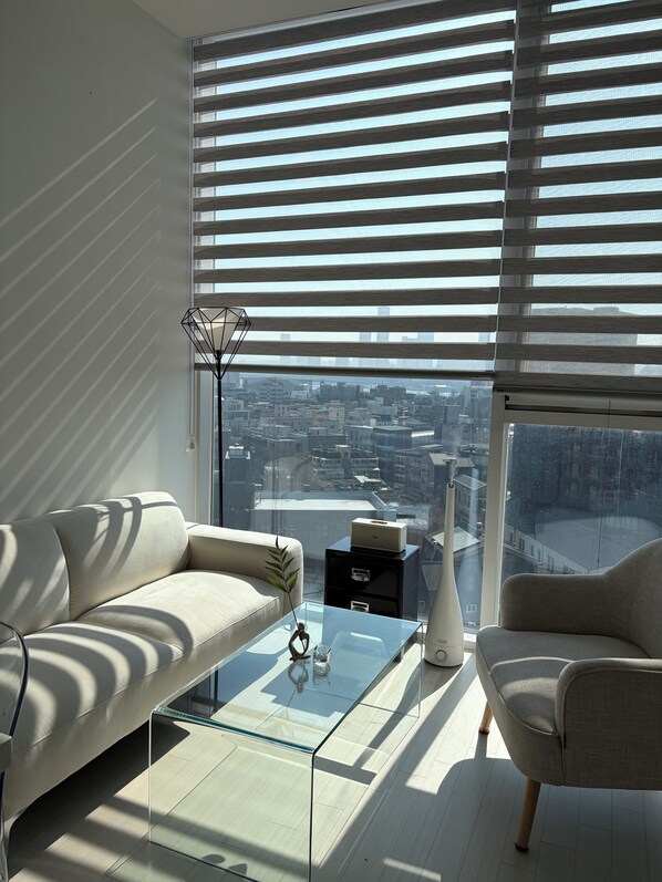 Un salon moderne comprenant un canapé, un fauteuil, une table basse en verre et une vue sur le paysage urbain, grâce à de grandes fenêtres équipées de stores.