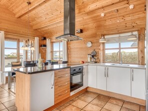 Eine moderne Küche mit Holzbalkendecke, einer zentralen Kochinsel, weißen Schränken und einem großen Fenster.