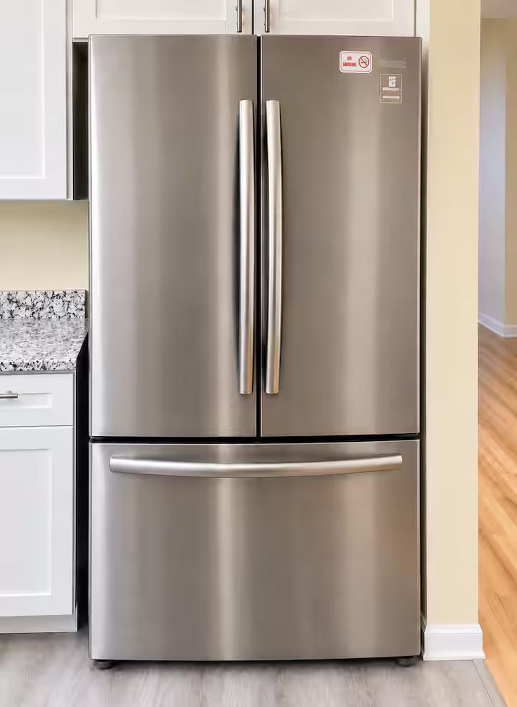 A stainless steel refrigerator with two doors and a freezer drawer, placed in a kitchen with white cabinets and a granite countertop.