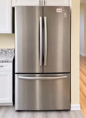 Un refrigerador de acero inoxidable con dos puertas y un cajón congelador, ubicado en una cocina con gabinetes blancos y una encimera de granito.