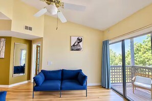 A living room with a blue sofa, a ceiling fan, a large window with a view, and a door leading to another room.