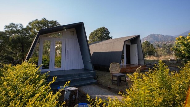 Two modern, tent-like structures with a flat roof, one with a visible door and steps leading to a porch area, and the other with a chair and table on the porch. - Dhauladhar View Village Resort (Dharamshala)