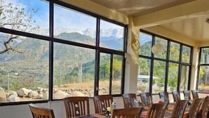 A dining area with a view of mountains, wooden chairs, a table with a colorful tablecloth, and a clock on the wall. - Dhauladhar View Village Resort (Dharamshala)