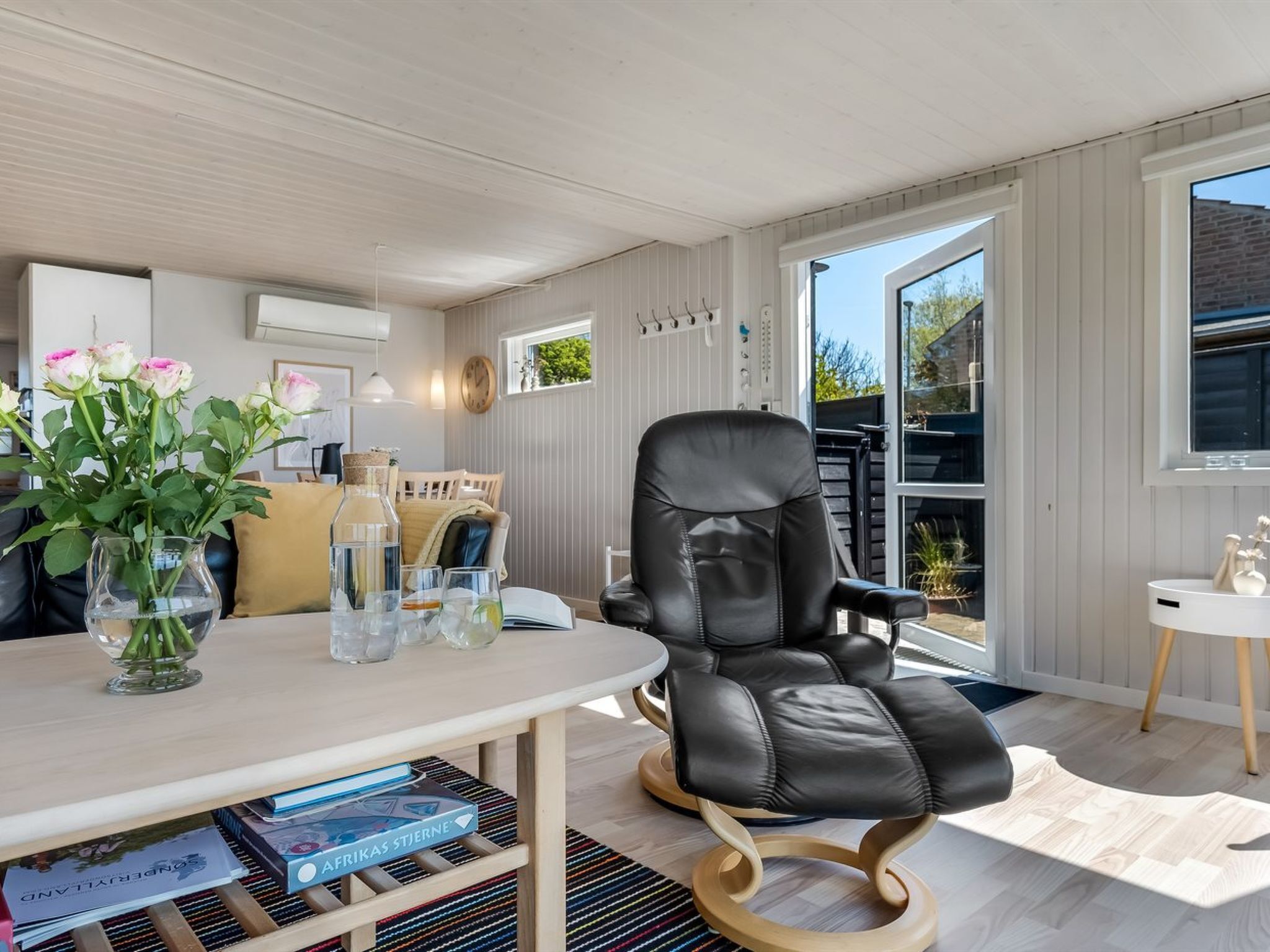 Un salon moderne avec un fauteuil en cuir, une table avec des livres et un vase de fleurs, et une vue sur l’extérieur.