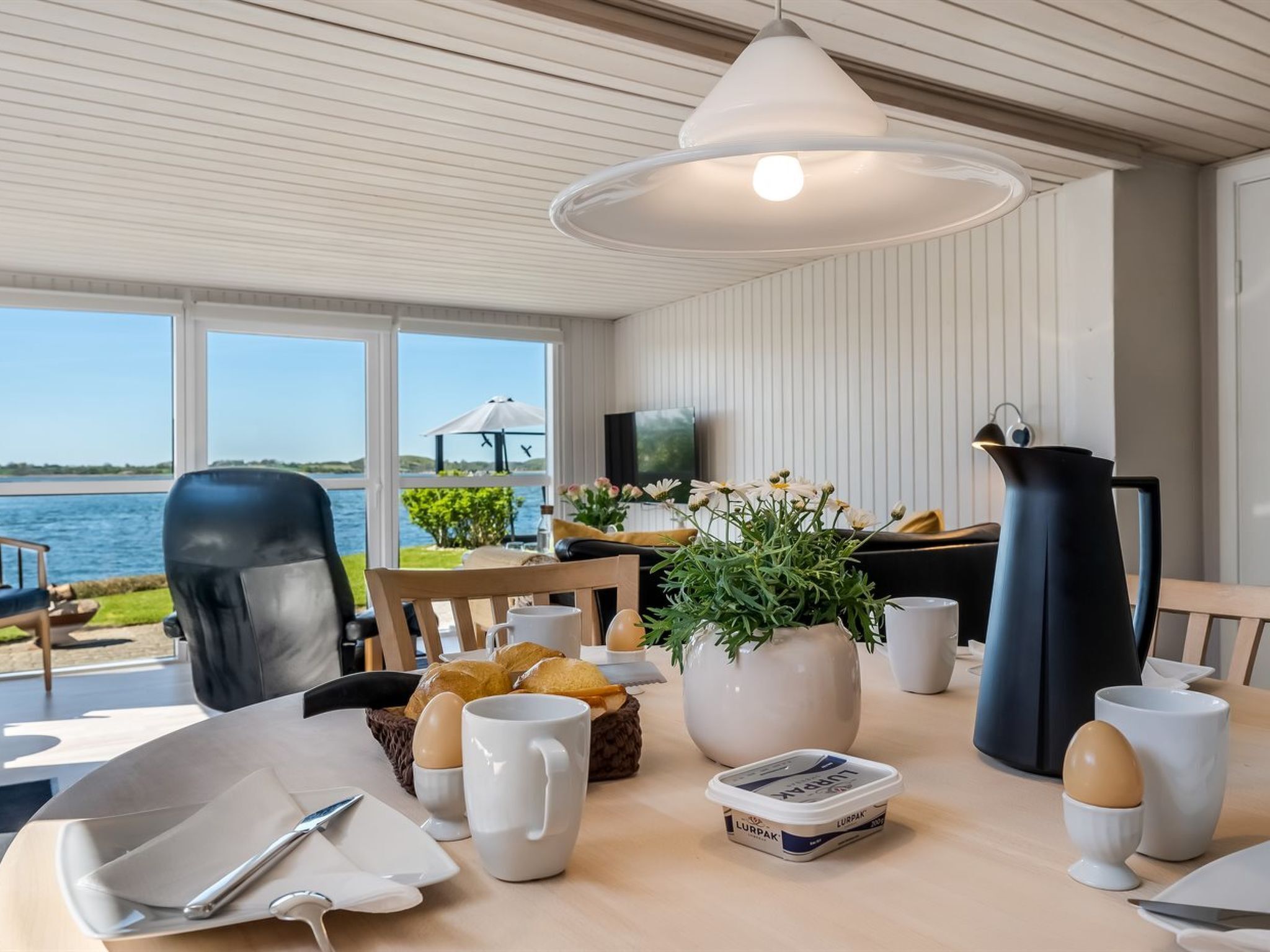 Een eetgedeelte met uitzicht op het water, een gedekte tafel met brood, eieren en een karaf, en een grote witte vaas met bloemen.