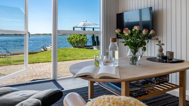 Ein Zimmer mit großem Fenster mit Blick auf ein Gewässer, einem Tisch mit Büchern und einer Blumenvase sowie einem Wand-Fernseher.