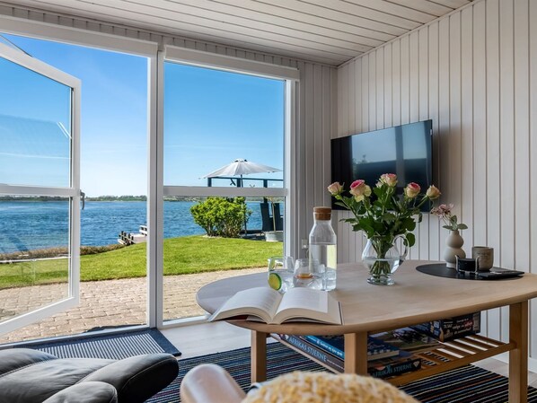 Ein Zimmer mit großem Fenster mit Blick auf ein Gewässer, einem Tisch mit Büchern und einer Blumenvase sowie einem Wand-Fernseher.