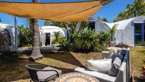 Un espace extérieur moderne aménagé avec une table et des chaises, entouré de plantes tropicales et protégé par une grande voile d’ombrage.