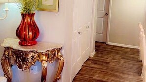 Ein Korridor mit Holzboden, einer weißen Tür, einem gerahmten Bild an der Wand und einer roten Vase mit Pflanzen auf einem Tisch.