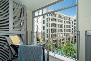 Ein Balkon mit Blick auf ein mehrstöckiges Gebäude, ein Tisch mit Flasche und Obst sowie ein Stuhl mit gemustertem Kissen.