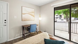 A room with a bed, a desk with a chair, a lamp, and a large window with a view of the street.