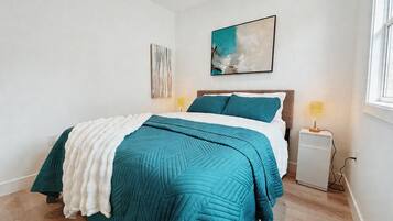 A bedroom with a bed, a bedside table, a lamp, a wall art piece, and a window.
