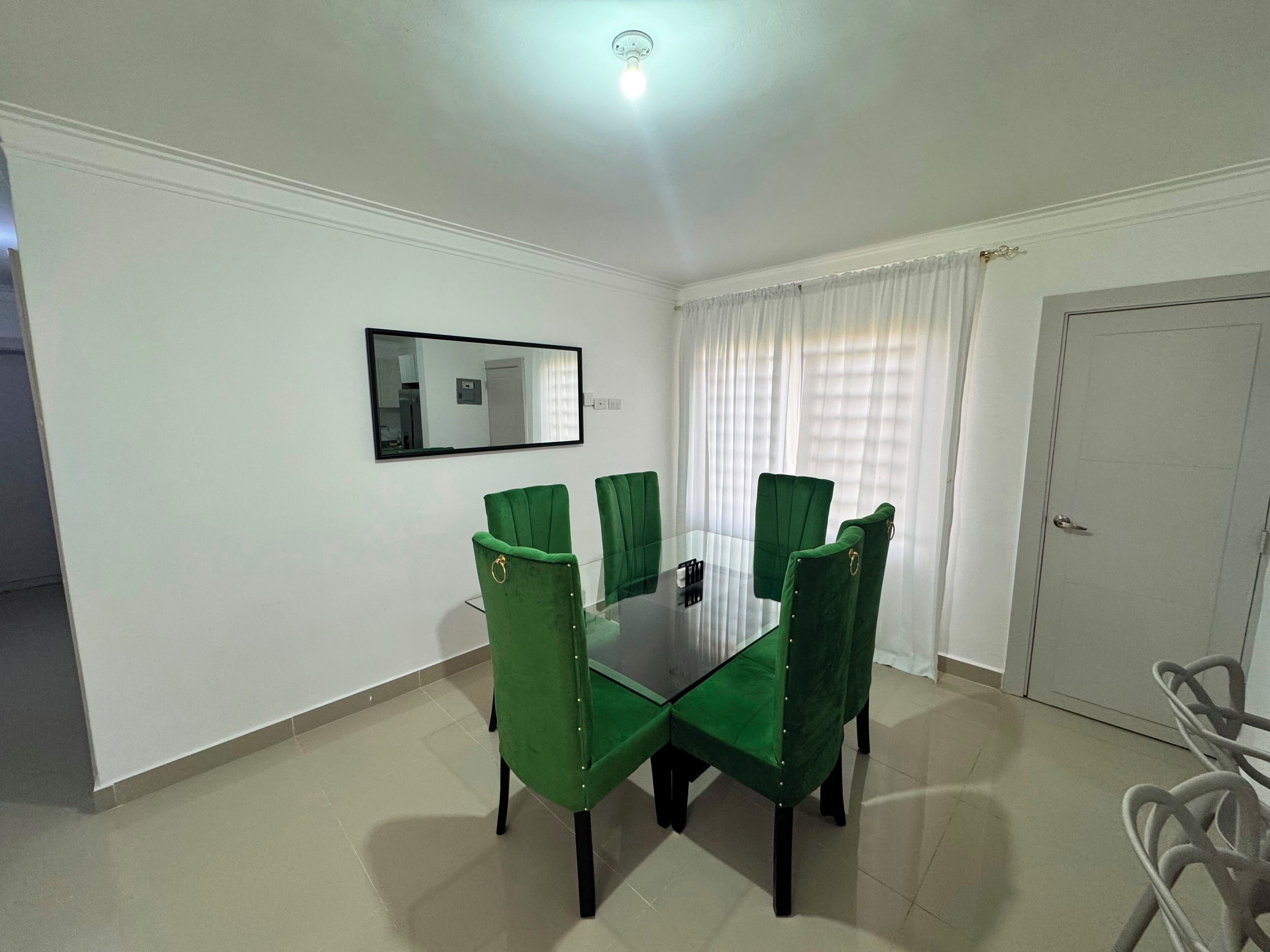 A dining area with a glass table surrounded by green upholstered chairs, a white door, and a mirror on the wall.