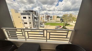 Ein Balkon mit zwei Korbstühlen und einem kleinen Tisch, mit Blick auf eine Straße mit Gebäuden und geparkten Autos.