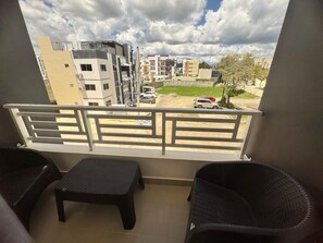 Un balcon avec deux fauteuils en osier et une petite table, donnant sur une rue bordée d’immeubles et de voitures garées.