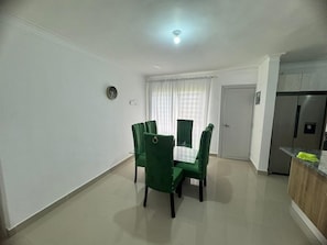 A dining area with a table and four chairs, a kitchen with a refrigerator, and a door leading to another room. - Luxury apartment  (San Francisco de Macorís)