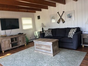 Un salon avec un canapé foncé, une table basse en bois, un téléviseur et un tableau encadré représentant deux pagaies croisées.
