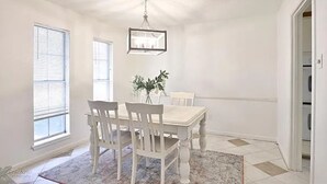 Un comedor con mesa y sillas blancas, lámpara colgante y ventana con persianas.