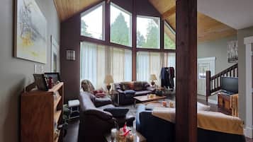 A cozy living room with a high ceiling, large windows, a fireplace, and a television.