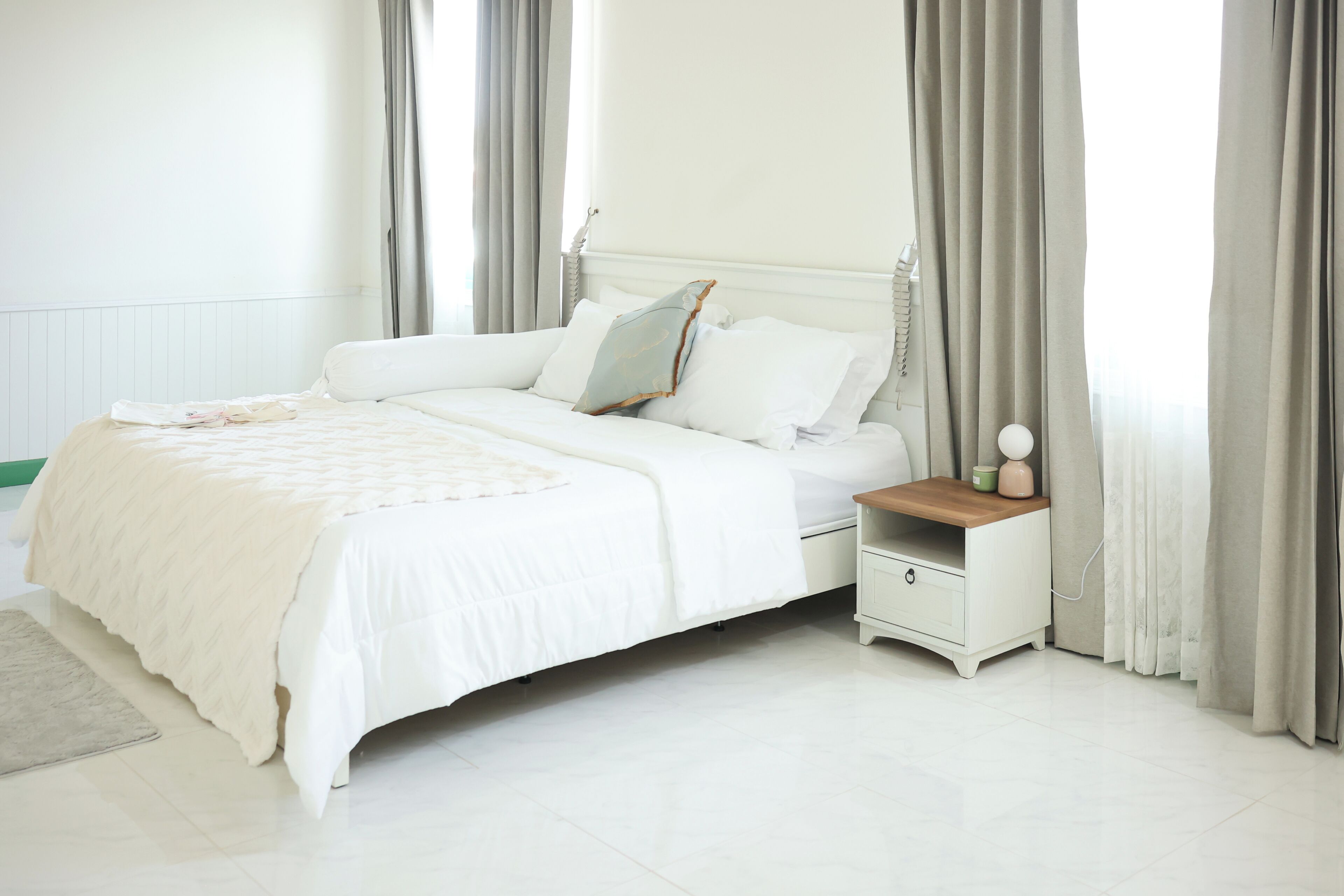 A neatly made bed with a white comforter and pillows, a bedside table with a lamp, and a nightstand with a cup and a small decorative item.