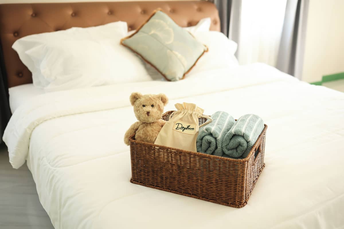 Un lit avec du linge de lit blanc, une tête de lit marron, un oreiller vert, un panier en osier contenant un ours en peluche, un sac et des serviettes enroulées.