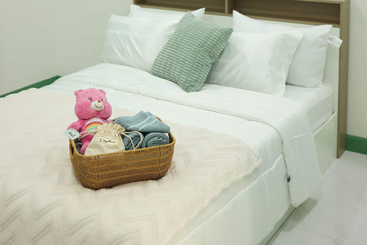 Un lit bien fait, avec un panier de vêtements et un ours en peluche, une table de chevet avec une horloge et un tableau encadré accroché au mur.