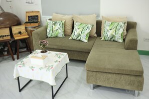 Un salon comprenant un canapé, deux fauteuils pouf, une petite table recouverte d’une nappe à motifs floraux et une boîte en bois sur laquelle est fixé un panneau d’ardoise.