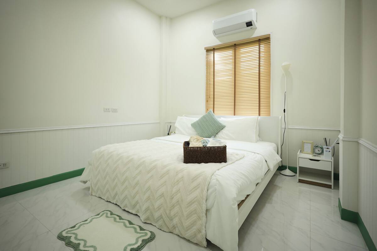 Une chambre à coucher comprenant un lit, une table de chevet, une commode, un climatiseur fixé au mur et une fenêtre avec des stores.